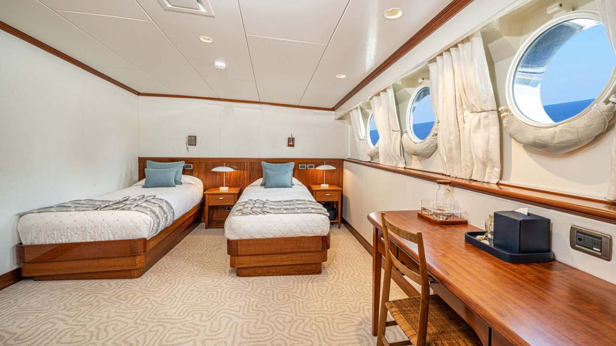 A well-furnished cabin with two twin beds, a wooden desk, and circular windows allowing natural light. Soft blue pillows on the beds complement the elegant décor.