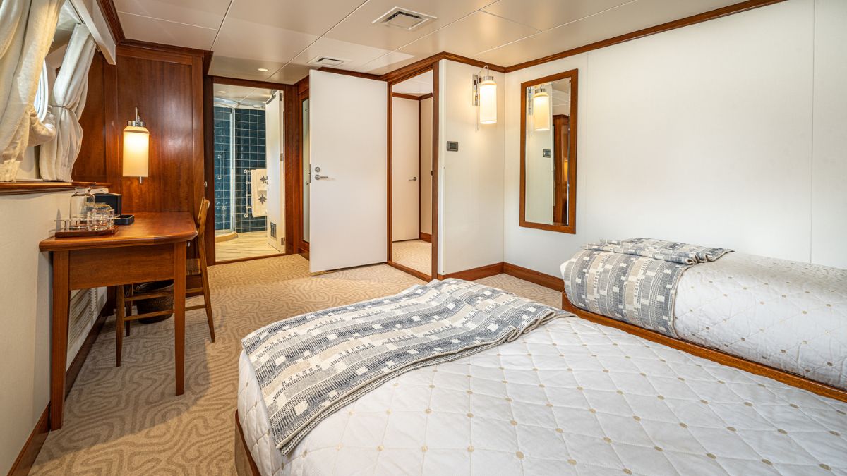 A well-lit cabin featuring two single beds with patterned throws, a wooden desk, and a full-length mirror. A bathroom entry is visible in the background.