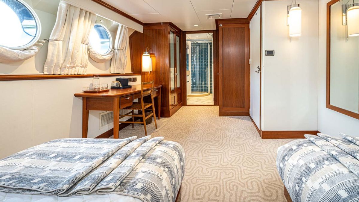 A luxurious yacht cabin featuring twin beds with patterned blankets, a wooden desk, and a cozy reading nook. Large round windows allow ample natural light, and an en-suite bathroom is visible in the background. The decor is elegant and nautical, with high-quality finishes.