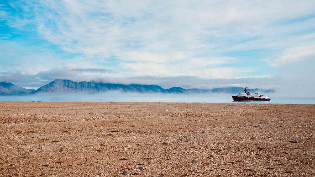 A large ship anchored near a rocky shoreline, with mist rolling over distant mountains under a partly cloudy sky.