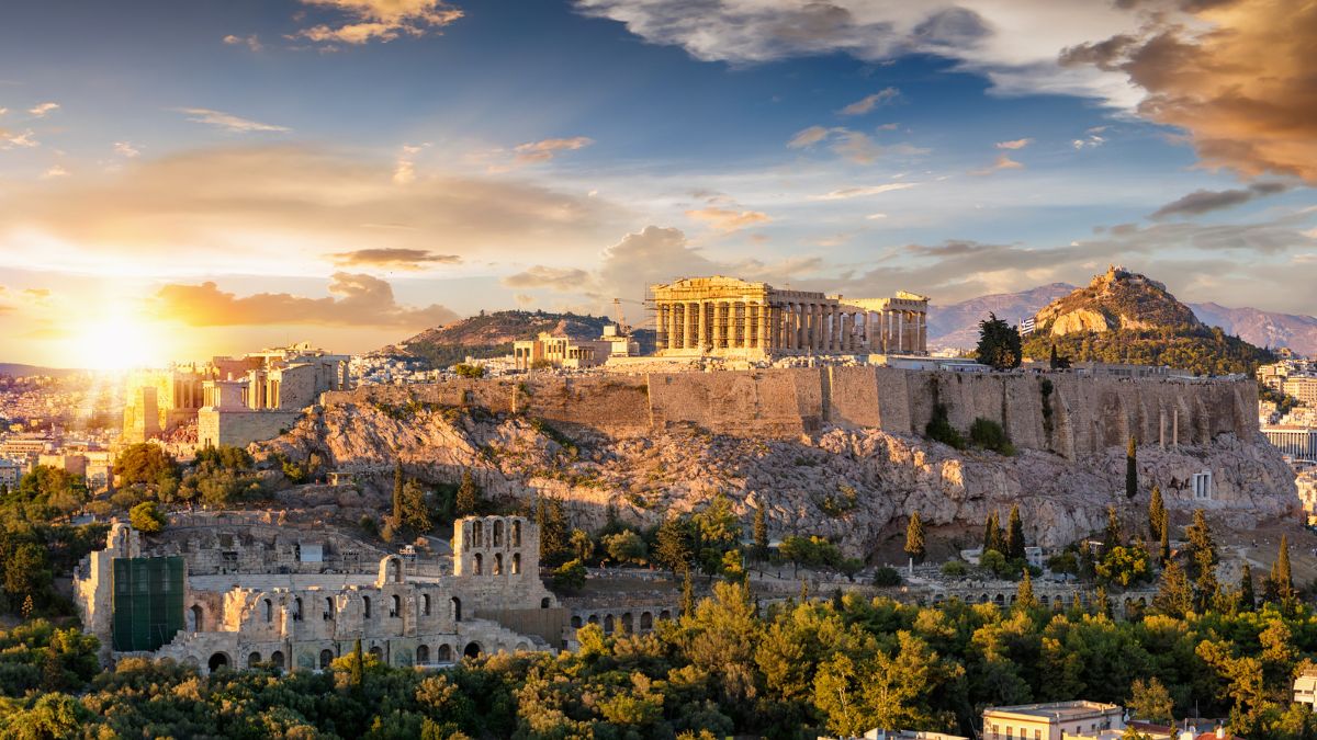 The Acropolis of Athens illuminated by a sunset, featuring the Parthenon atop a rocky hill surrounded by ancient ruins and lush greenery.