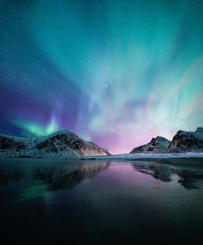 Northern Lights in Norway illuminating the sky with shades of green, purple, and blue over snow-capped mountains, reflected in a calm body of water below. Stars twinkle in the sky.
