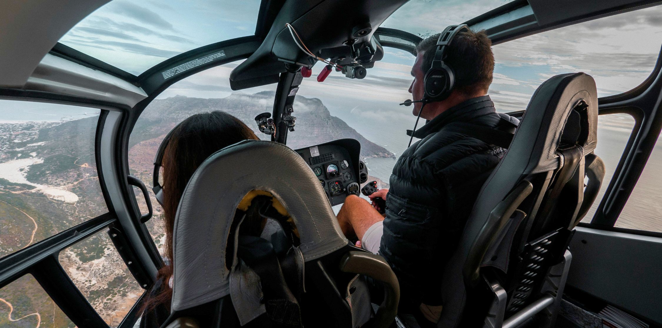 A helicopter cockpit view shows a pilot wearing a headset while flying over a coastal landscape. The passenger, partially visible, is seated behind the pilot, with rugged terrain and sea below.