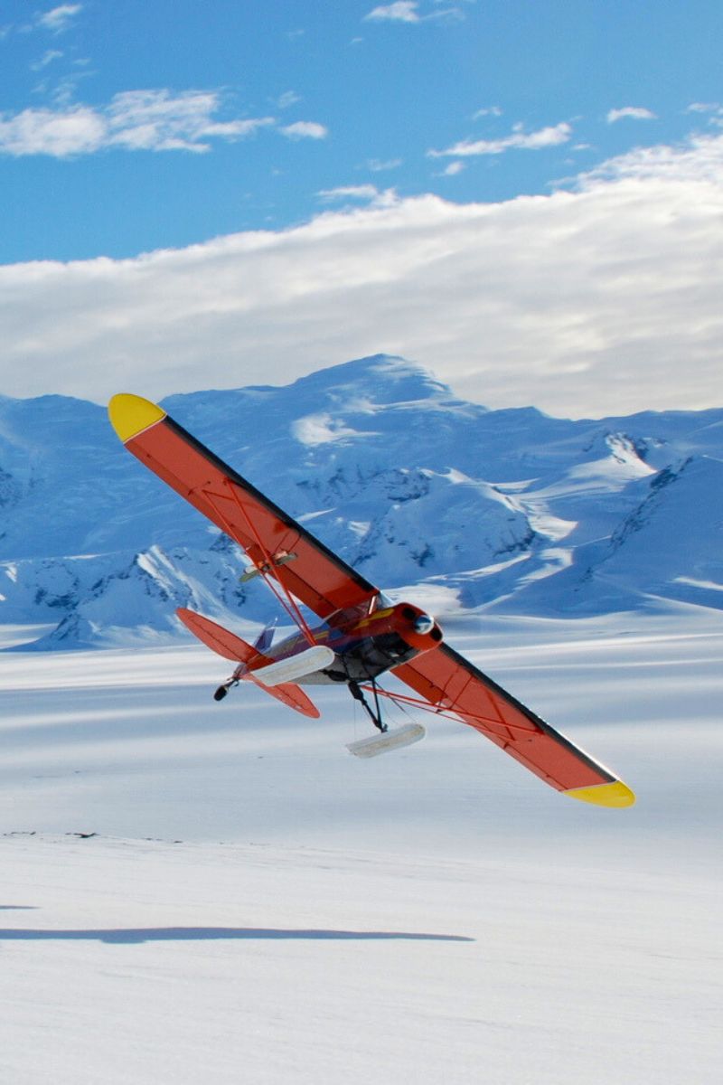 An orange and yellow plane flying low over a snowy landscape with mountains in the background under a partly cloudy sky.