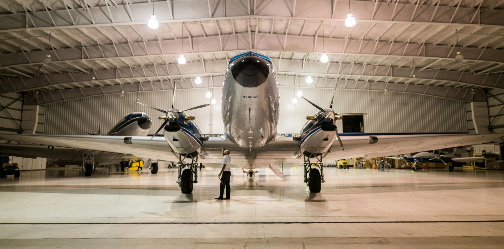 A vintage aircraft is parked in a spacious hangar, with a person standing in front of it, admiring the airplane's sleek design and polished surface. The hangar's high ceiling and bright lighting highlight the aircraft's features.