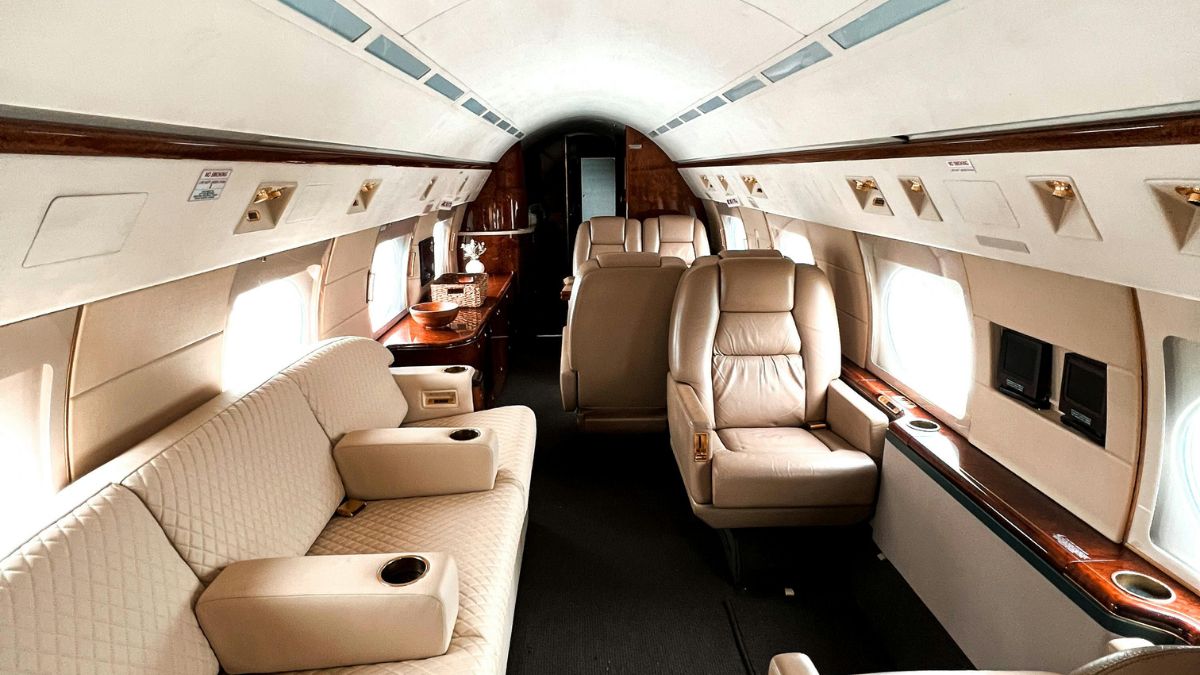 Luxurious interior of a private jet featuring a spacious seating area with cream-colored leather sofas and armchairs, elegant wood accents, and large windows allowing natural light.