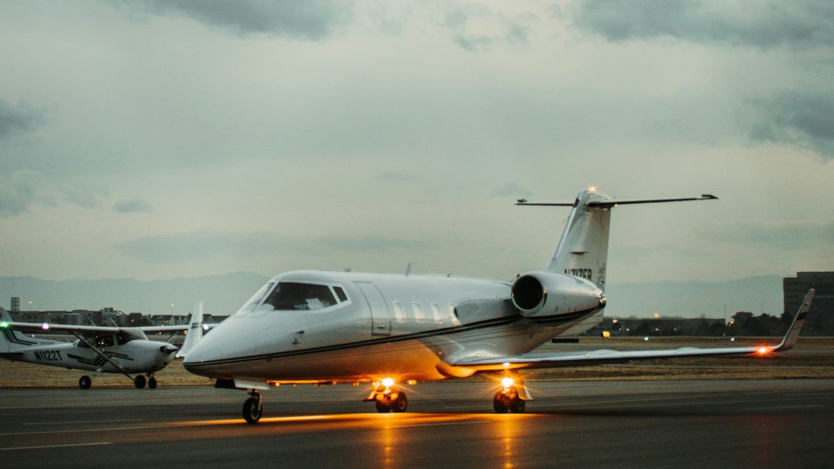 A sleek business jet taxiing on the runway, with its landing lights illuminated. In the background, another small aircraft is parked. Dark, cloudy sky overhead.