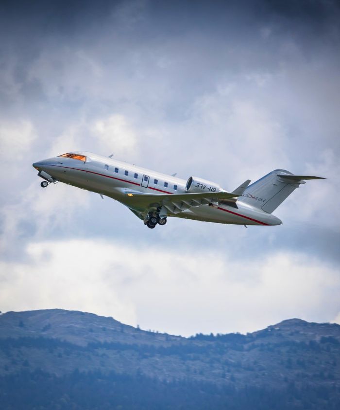 A silver private jet taking off against a backdrop of cloudy skies and mountains.