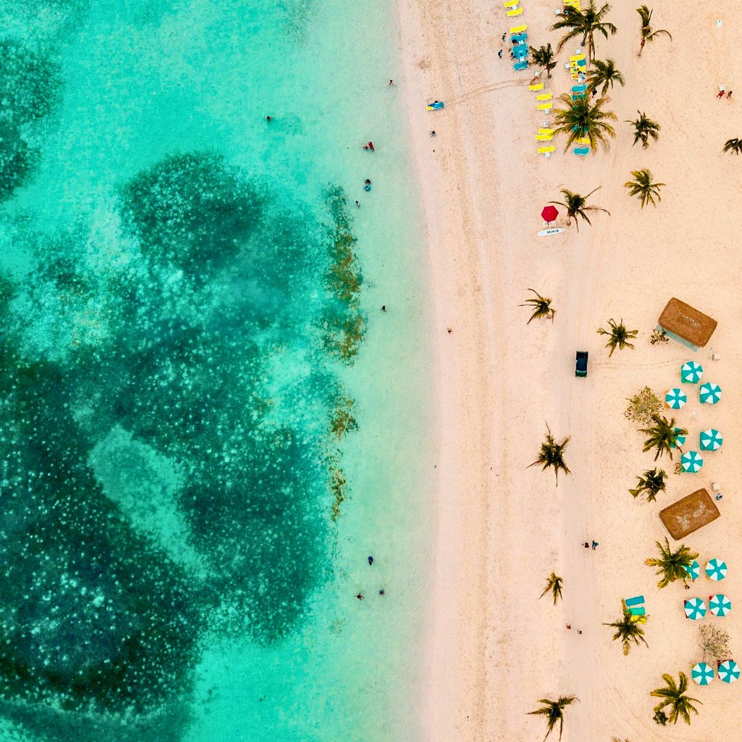 Aerial view of a tropical beach with soft white sand and turquoise waters. Palm trees line the shore, with colorful beach umbrellas and loungers visible. Some people are swimming in the clear water, while others relax on the sand. Coral formations can be seen beneath the water's surface.