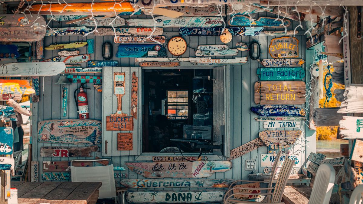 A colorful shack adorned with various wooden signs and messages, displaying a mix of names, places, and humorous phrases, against a sunny backdrop.