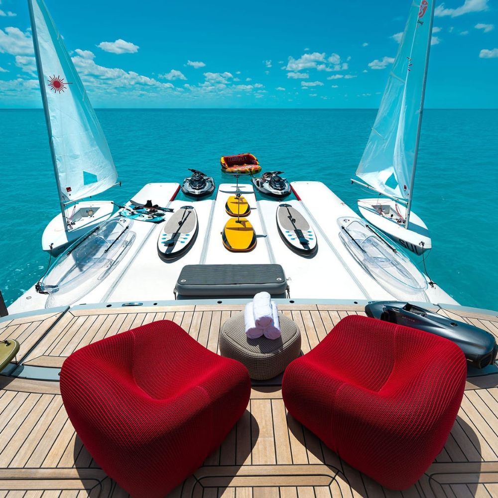 Two red chairs sit on a sleek yacht deck, overlooking turquoise waters. Several small sailboats and watercraft are neatly arranged on the deck, with a bright blue sky and a few clouds visible in the background.