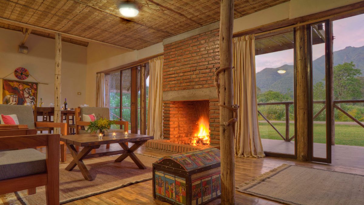 Cozy living room with a brick fireplace, wooden furniture, and large windows showcasing a mountain view. Warm light from the fire creates a welcoming atmosphere.