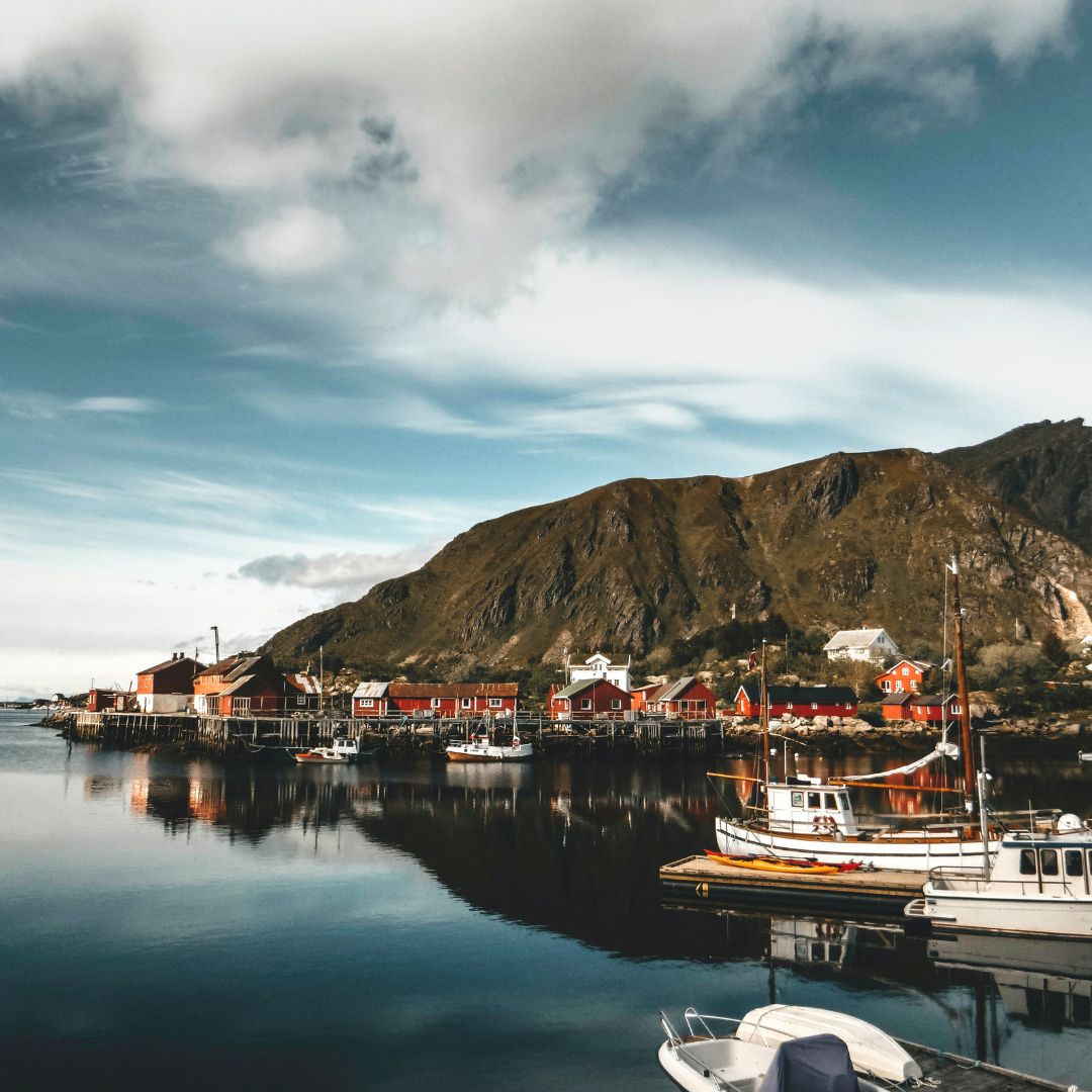 A tranquil harbor scene featuring colorful buildings along the waterfront, surrounded by a mountainous landscape. Boats are moored in the still water reflecting the sky.