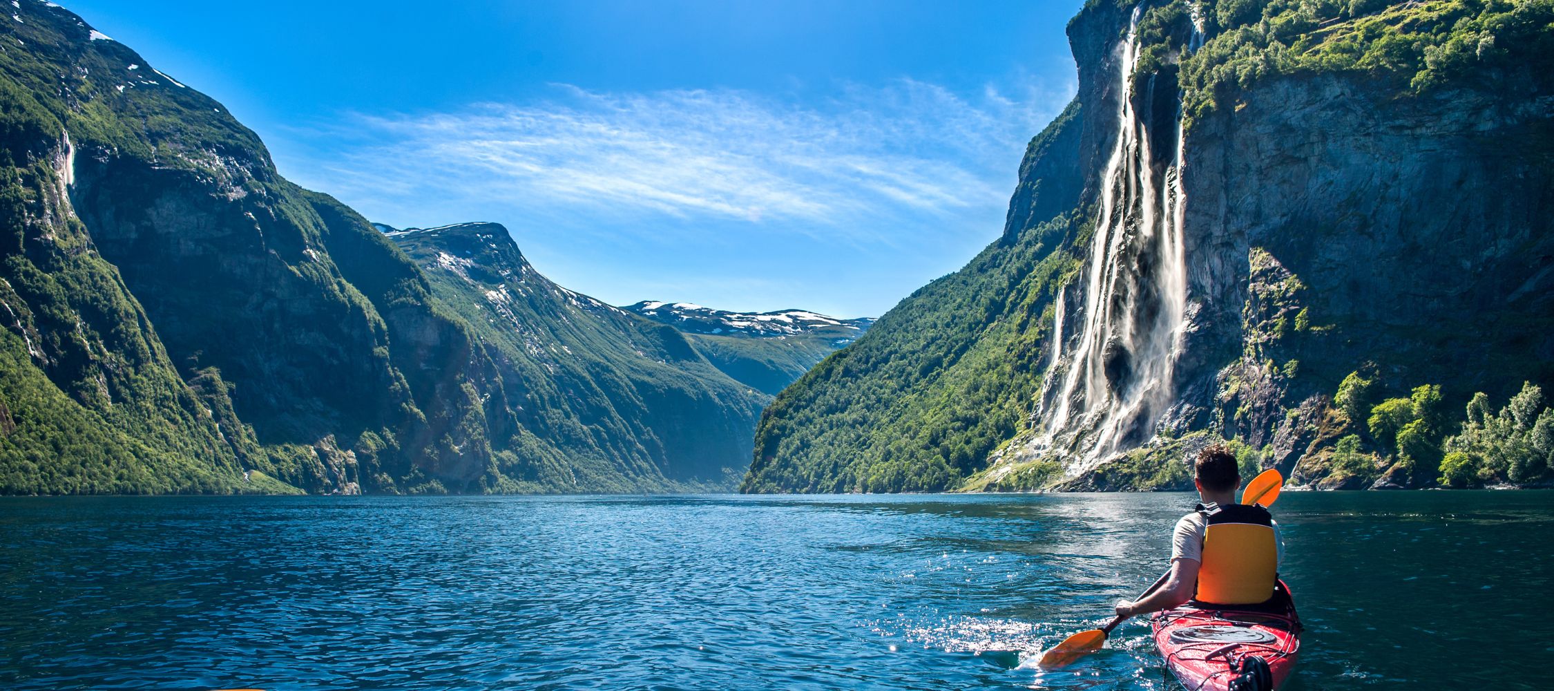 A kayaker paddles on a serene fjord, surrounded by towering cliffs and lush greenery. A breathtaking waterfall cascades down from the mountains in the background under a clear blue sky.