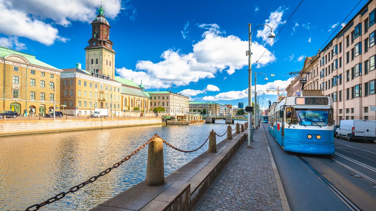 Tram passing along a waterfront street with historic buildings and a clock tower under a bright blue sky with fluffy clouds.