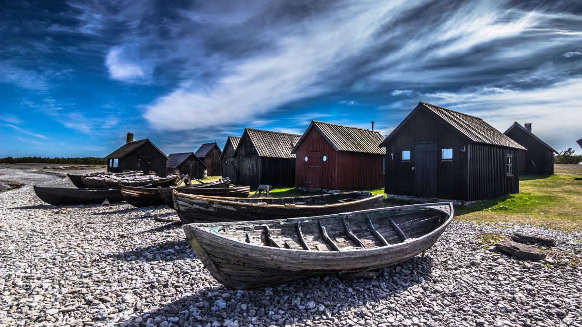 Abandoned wooden boats on a rocky shore, with a line of black and red fishing huts under a dramatic blue sky with wispy clouds.