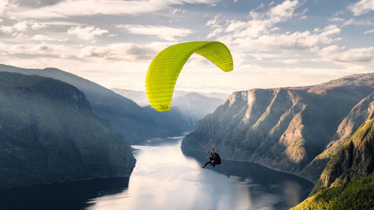 A person paraglides with a bright yellow wing above a serene fjord, surrounded by lush green mountains under a partly cloudy sky.