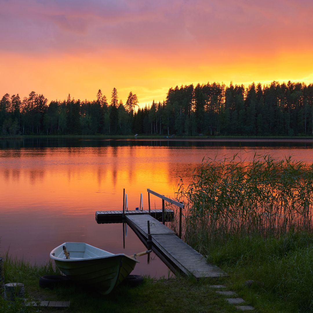 A tranquil lake at sunset with vivid orange and purple skies reflecting on the water. A small rowboat rests on the shore near a wooden dock, surrounded by lush greenery and tall reeds.