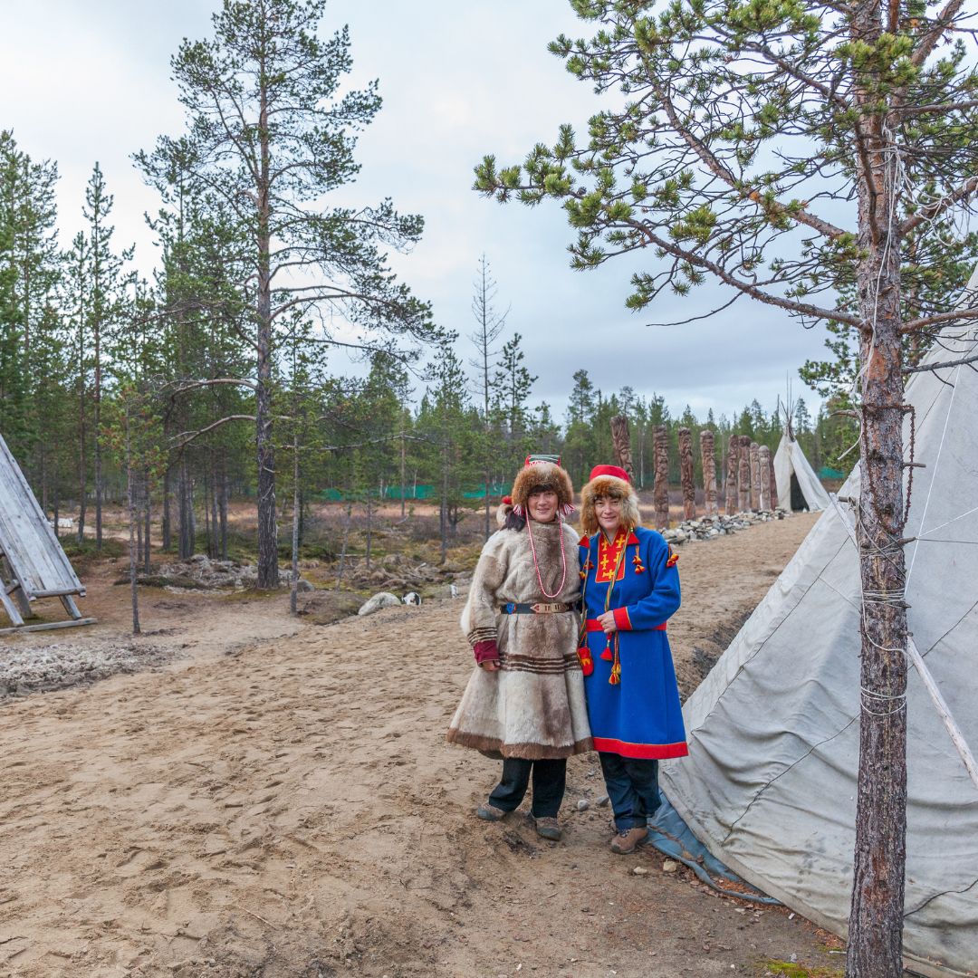 Two individuals dressed in traditional Sami clothing stand together in a sandy area surrounded by pine trees. They are near teepee-like structures, with one person wearing a fur coat and the other in a colorful outfit.