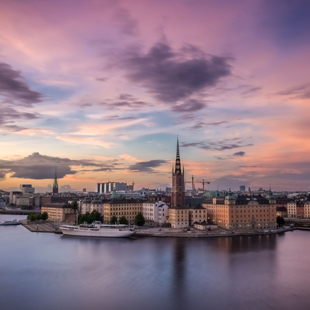 A picturesque view of Stockholm at sunset, showcasing the skyline with the iconic spires of Gamla Stan and modern buildings. The water reflects the vibrant colors of the sky, creating a serene atmosphere.