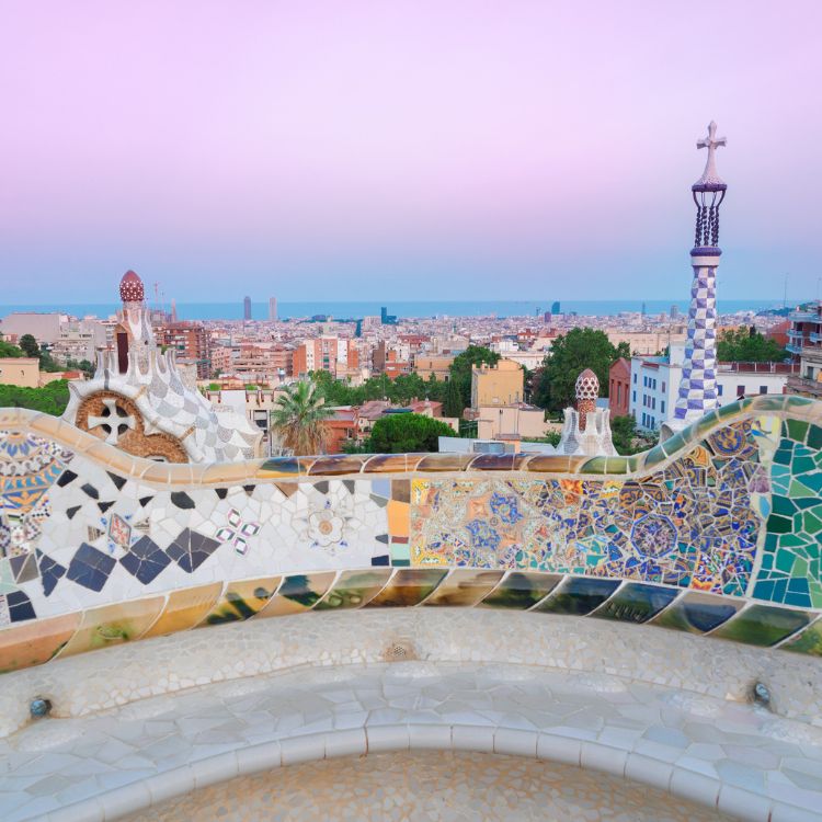 A panoramic view of Barcelona from Park Güell, showcasing colorful mosaic tile work, a distant skyline, and a serene pastel sky at dawn.