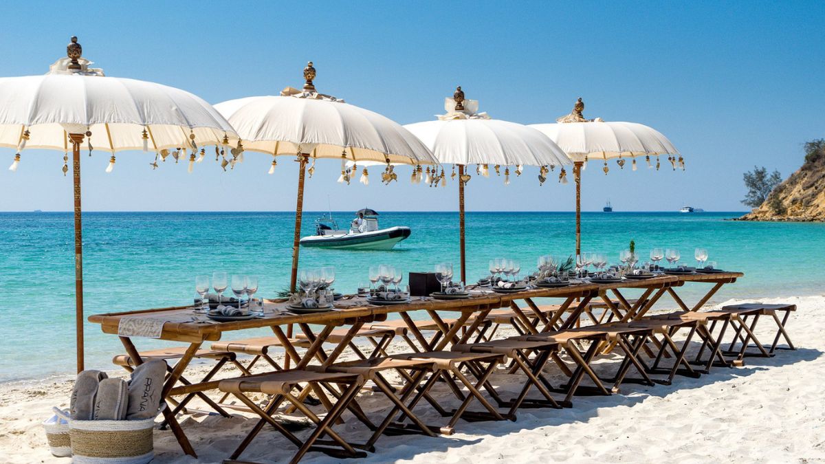 A beautifully arranged beach dining setup featuring a long wooden table with elegant tableware, shaded by large white umbrellas adorned with decorative elements. The turquoise ocean and a small boat are visible in the background, providing a serene coastal ambiance.