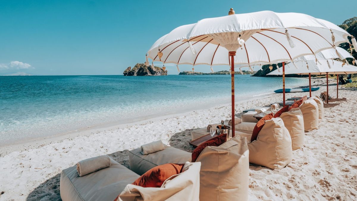 Sun loungers with cushions and large umbrellas line a sandy beach beside clear blue water, with rocky formations visible in the distance.