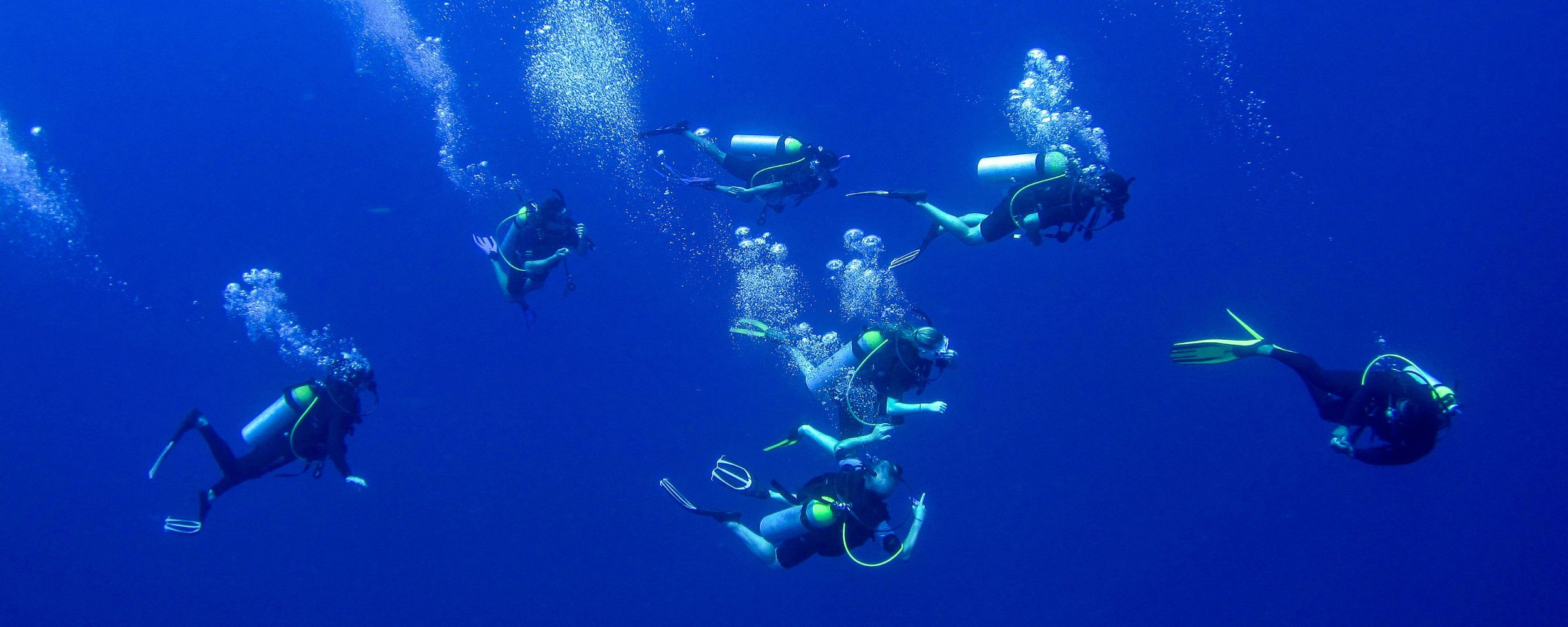 A group of eight divers underwater, surrounded by deep blue water and bubbles. They are wearing scuba gear and fins, exploring the ocean environment.