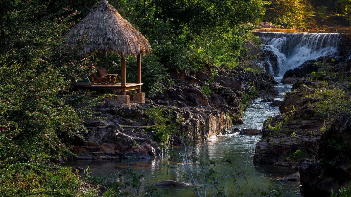 A thatched gazebo with wooden chairs sits by a serene river, surrounded by lush greenery and rocky terrain. A waterfall cascades in the background, illuminated by soft sunlight.