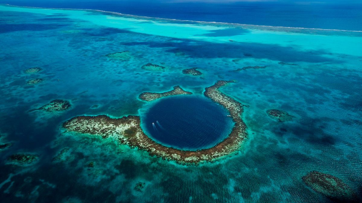 Aerial view of a circular blue hole surrounded by vibrant turquoise waters, with patches of coral and small islands scattered around.