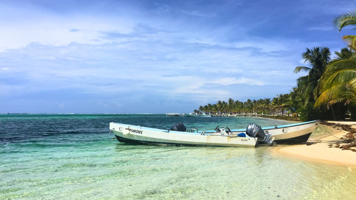A serene beach scene featuring a small white boat anchored near the shore. Clear turquoise waters gently lap against a sandy beach, with palm trees lining the coast in the background under a blue sky with scattered clouds.