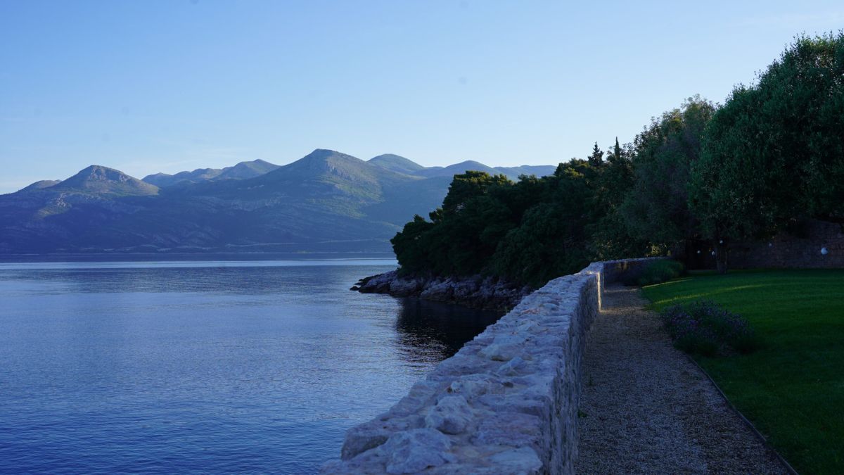 A serene coastal scene with a stone wall lining the water's edge. In the background, mountains rise against a clear blue sky, reflecting on the calm water. Lush greenery and trees border the pathway, creating a tranquil atmosphere.