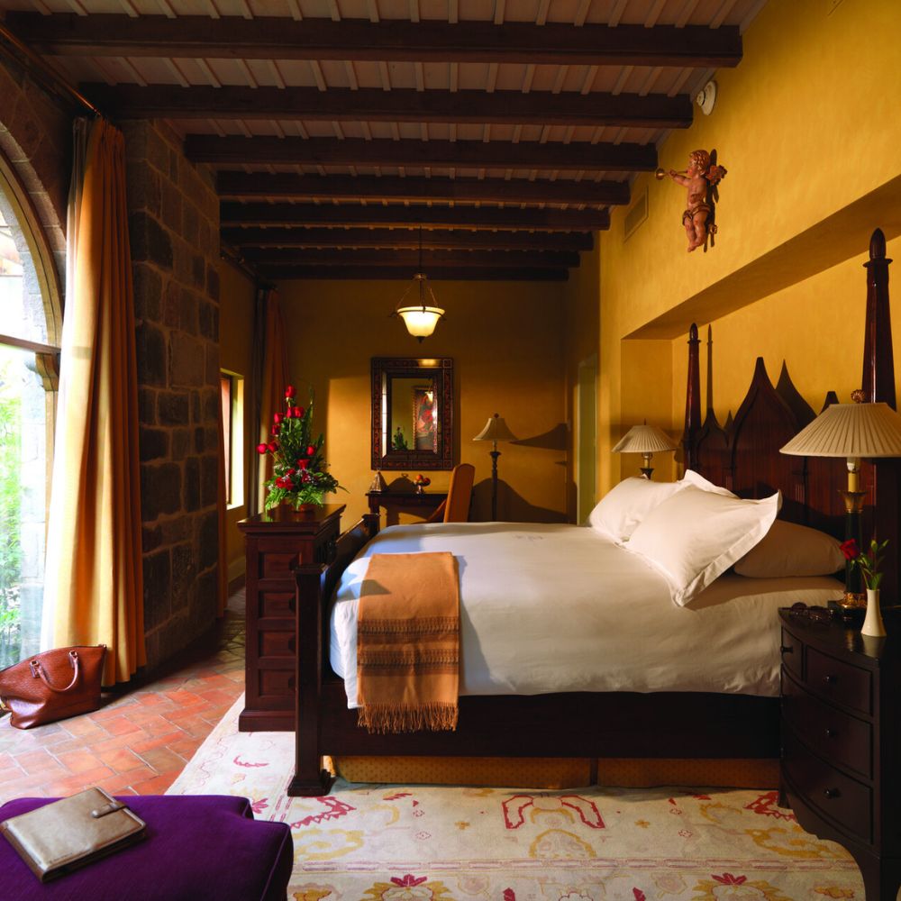 Cozy hotel room featuring a wooden bed with white linens, warm yellow walls, and rustic wooden beams. Decorative elements include a mirror, lamps, and a bouquet of flowers on a bedside table. A soft rug and a stylish handbag add to the inviting atmosphere.