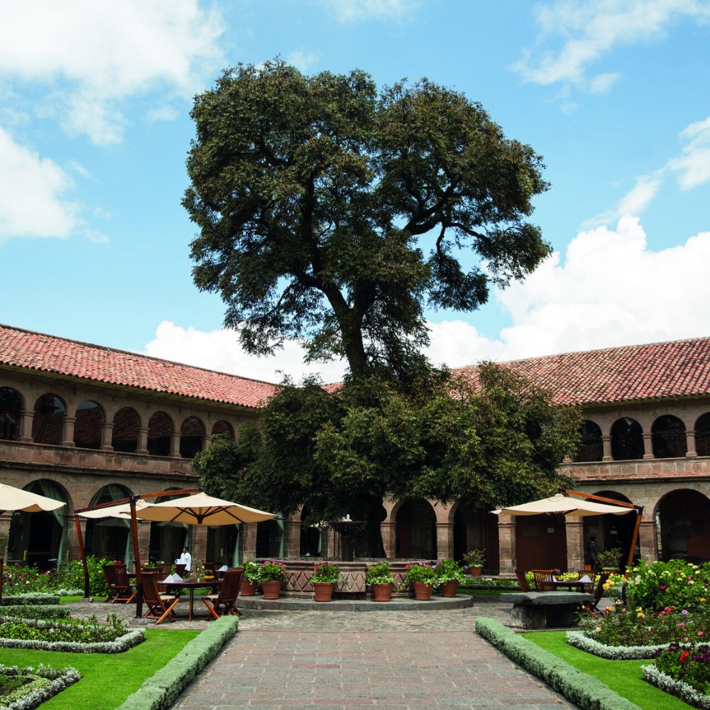 A serene courtyard surrounded by lush greenery, featuring a large tree in the center. Wooden tables and chairs are set under umbrellas, with blooming flowers and stone pathways adding to the tranquil atmosphere. The sky is partly cloudy, enhancing the peaceful scene.