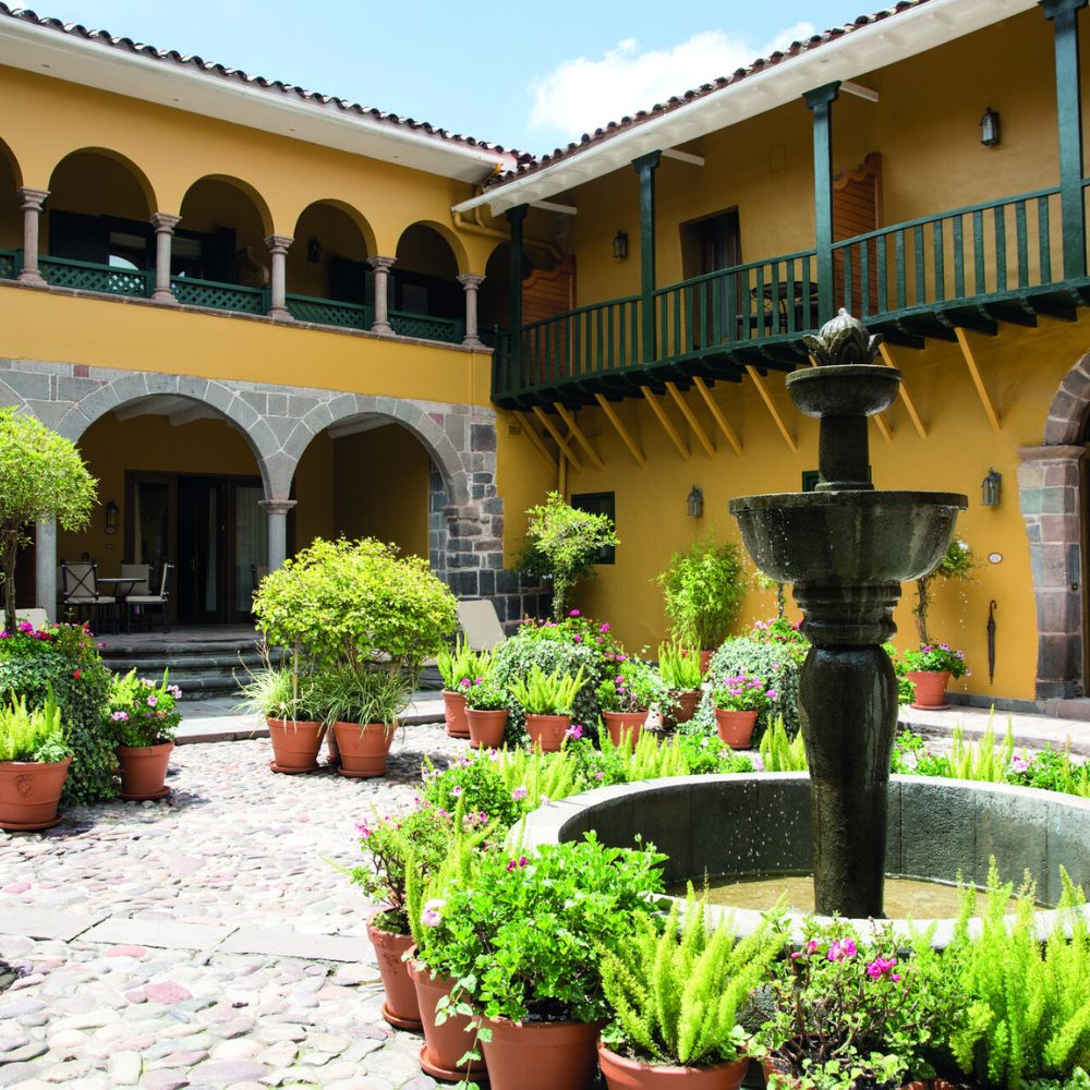 A charming courtyard featuring a decorative fountain surrounded by lush green plants and pots. Arched entryways lead into a warm, colorful building with a balcony above. The cobblestone floor adds to the rustic ambiance.