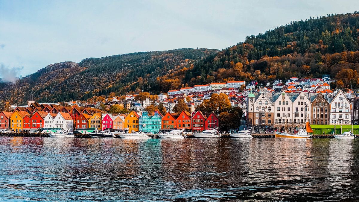 Colorful, historic wooden buildings line a waterfront, reflecting in calm waters. Surrounding hills are adorned with autumn foliage. Several boats are docked at the shore.