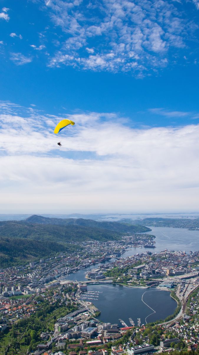 A paraglider with a yellow canopy soars over a picturesque coastal city, surrounded by green hills and water. The sky is bright with scattered clouds.