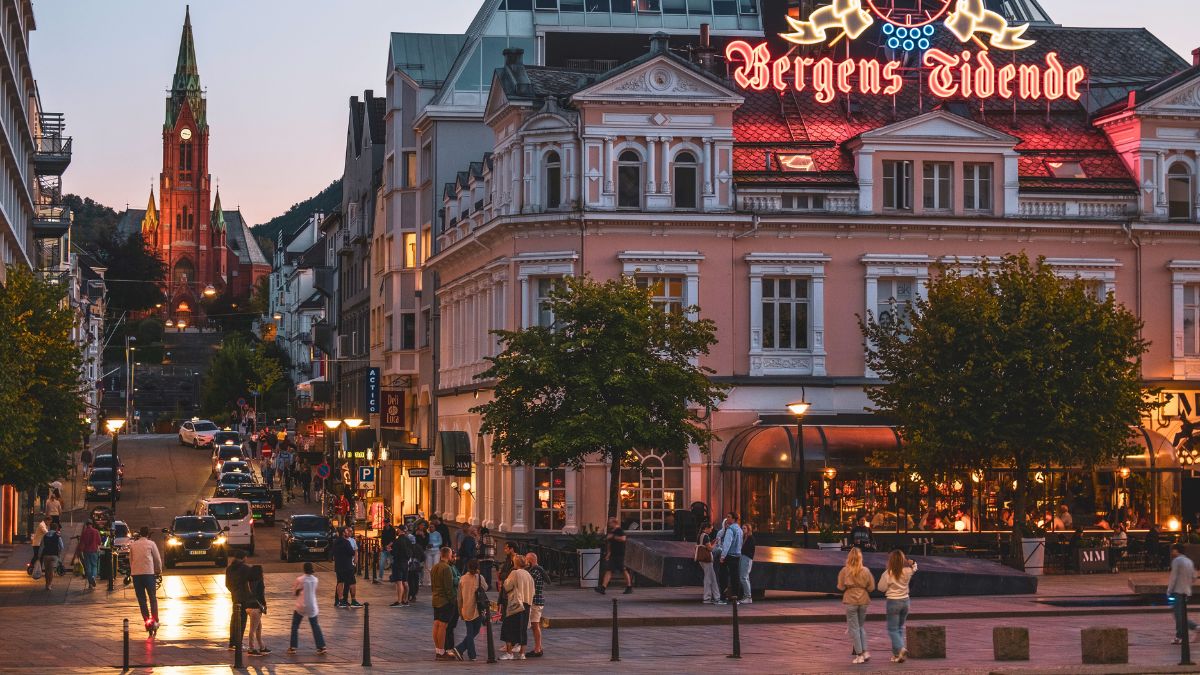 A bustling street scene in Bergen, Norway, with people walking in front of a restaurant and the lit-up "Bergens Tidende" sign above. A hill in the background features a tall, red church illuminated by evening light.