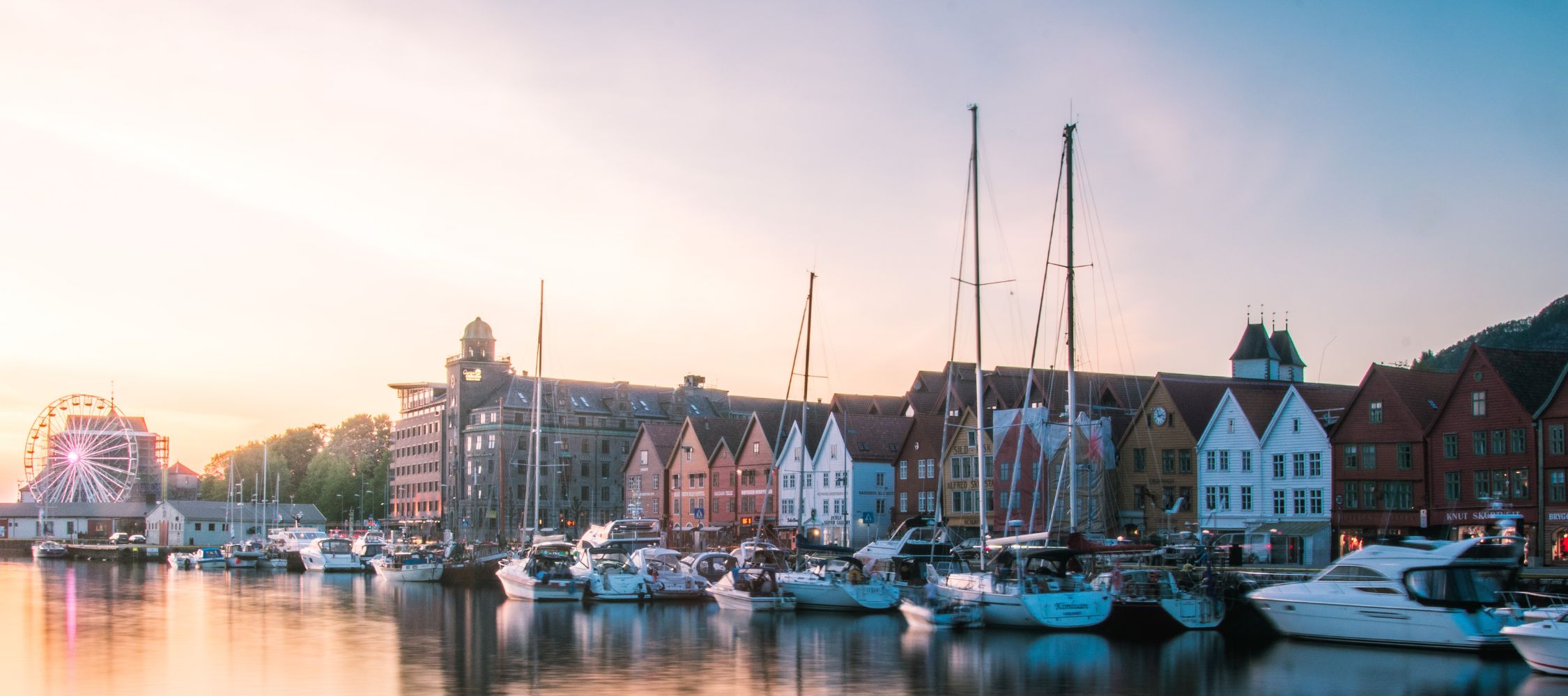 A harbor at sunset featuring several boats docked alongside colorful waterfront buildings. A Ferris wheel is visible in the background, adding to the scenic view.