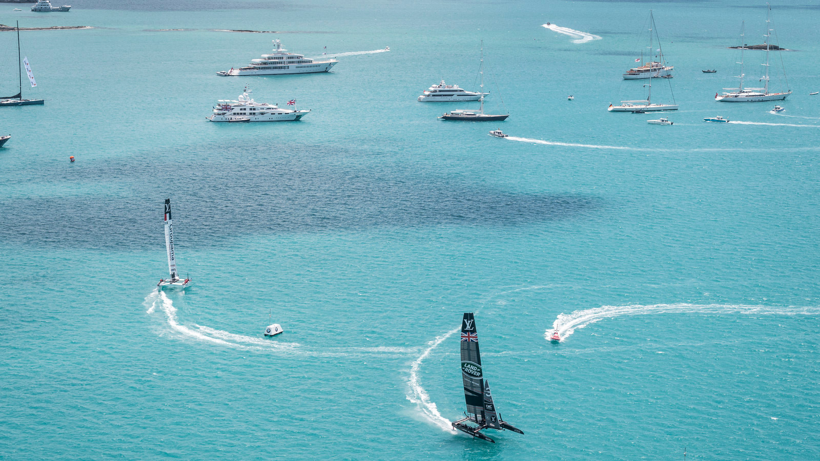 Sailboats and yachts navigate the turquoise waters, with some boats leaving distinct wake patterns as they move. A sleek racing catamaran can be seen in the foreground, competing with other vessels in the scenic seascape.
