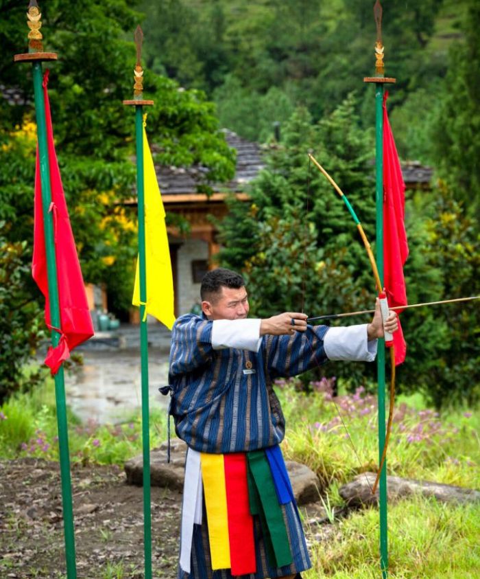 A man in traditional Bhutanese attire stands poised to shoot an arrow, with colorful flags and greenery in the background.