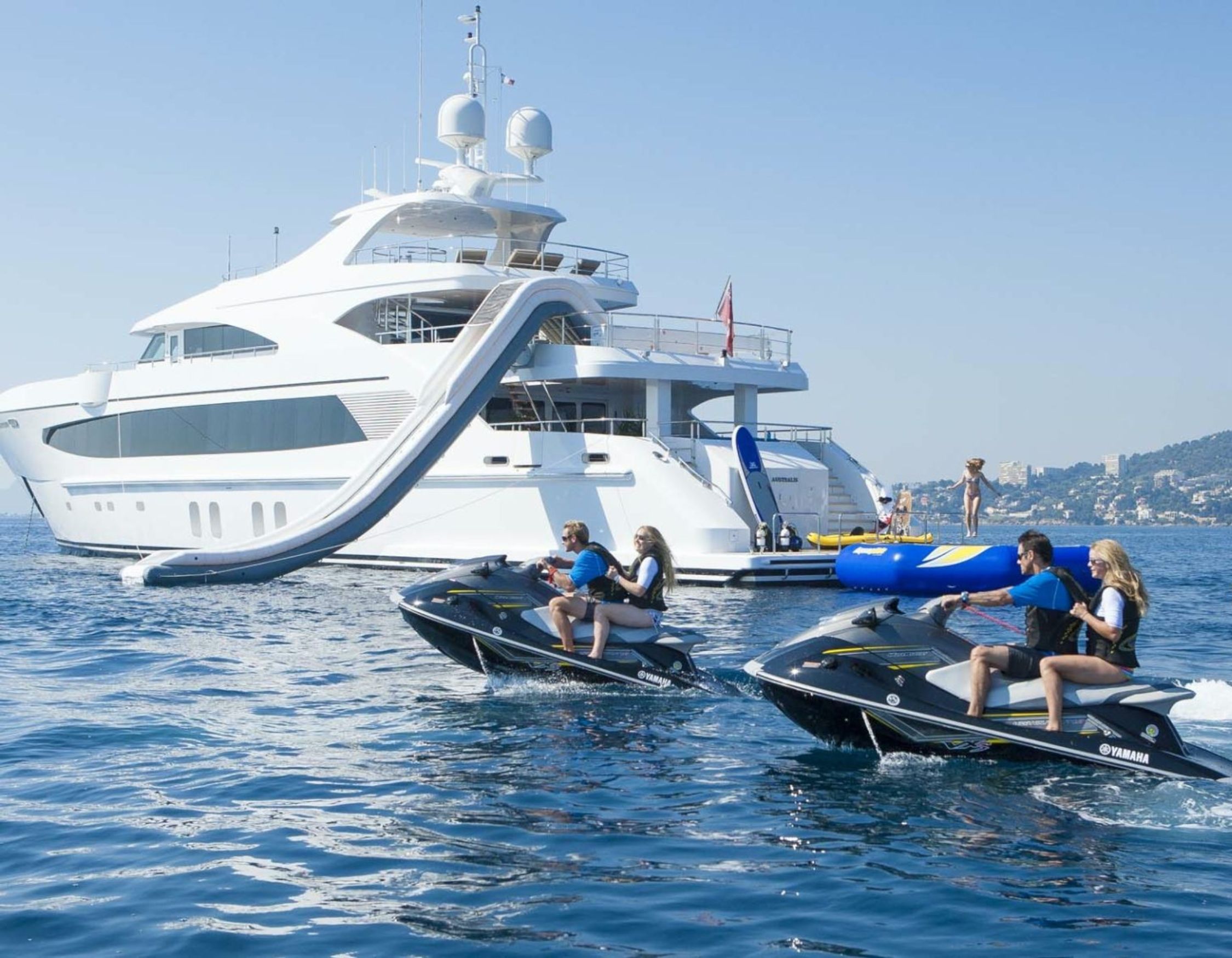 Two jet skis with riders speeding across the water, accompanied by a large luxury yacht in the background featuring a waterslide. The scene is set in a sunny, clear day by the coast.