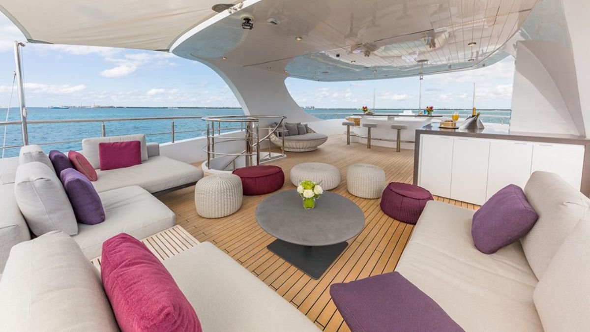 A luxurious outdoor lounge area on a yacht features comfortable seating with pillows, a round table, and views of the ocean. Elegant décor includes colorful cushions and a wooden deck.