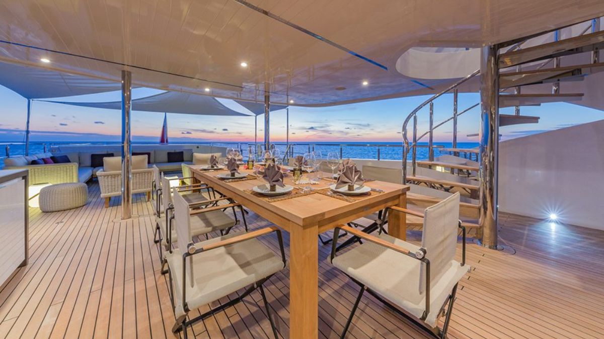 A beautifully set outdoor dining area on a yacht, featuring an elegant wooden table surrounded by chairs, with neatly arranged tableware and napkins. The scene is illuminated by soft lighting, with a stunning sunset view over the water in the background.