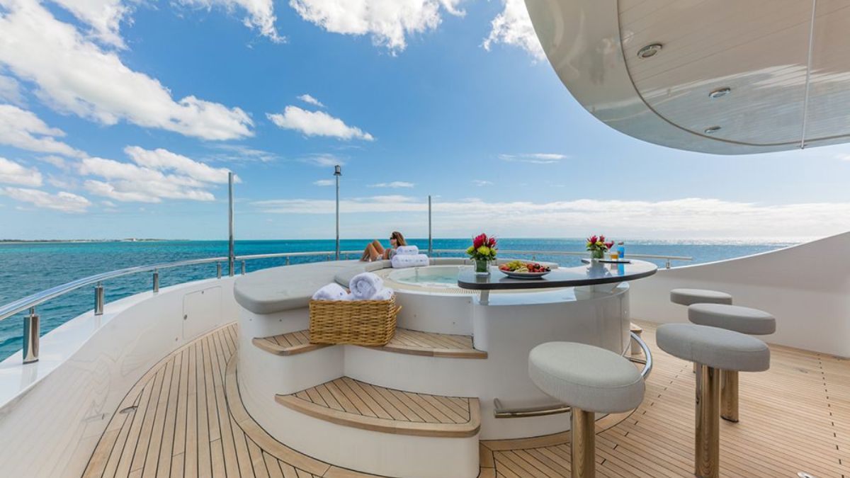 A luxurious yacht deck with a stylish round bar area and comfortable stools. A person relaxes nearby, surrounded by a stunning ocean view and blue sky. A basket of towels and a platter of fruit are neatly arranged on the bar.