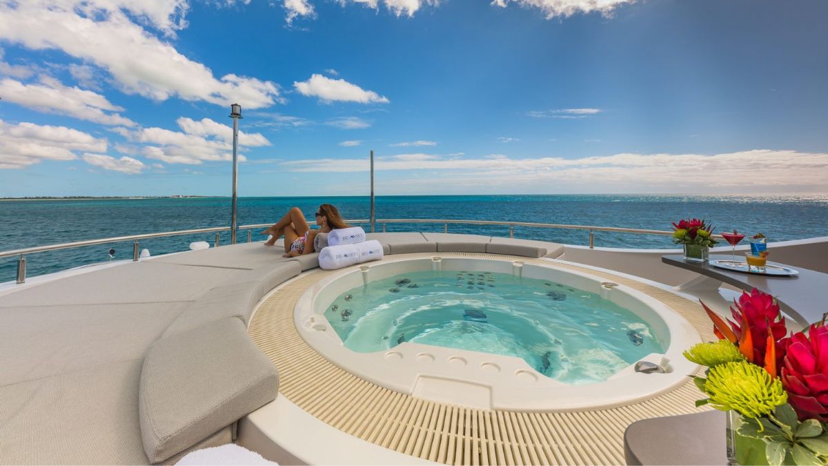A woman relaxes beside a hot tub on the deck of a luxurious yacht, overlooking a sparkling blue sea under a partly cloudy sky. Bright flowers are arranged on a nearby table.