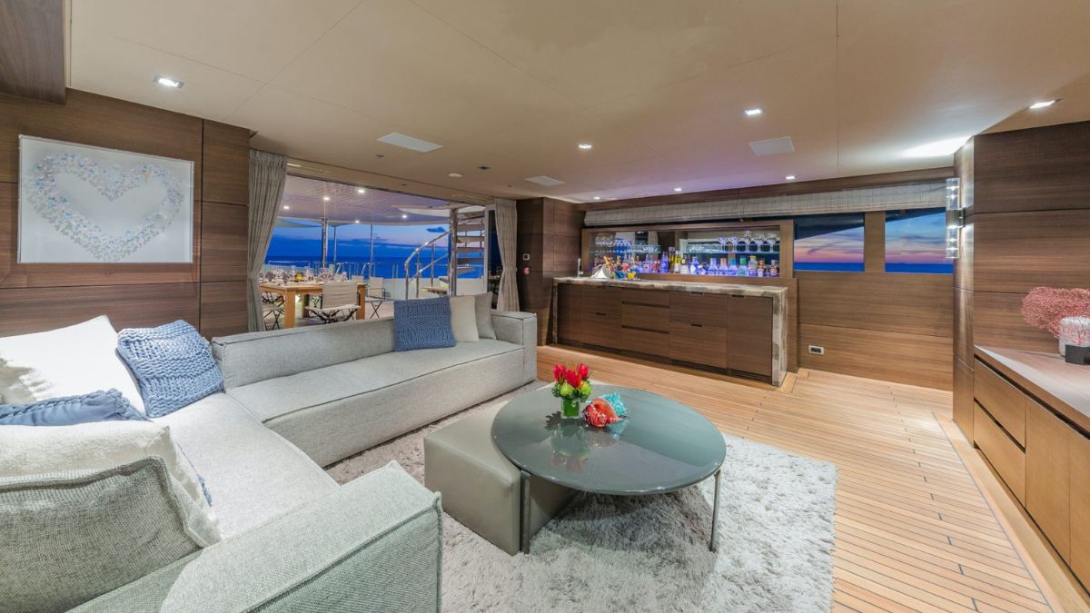 Luxurious yacht interior featuring a spacious lounge area with a comfortable gray sectional sofa, a round glass coffee table adorned with flowers, and a stylish bar showcase. Large windows provide stunning ocean views during twilight.