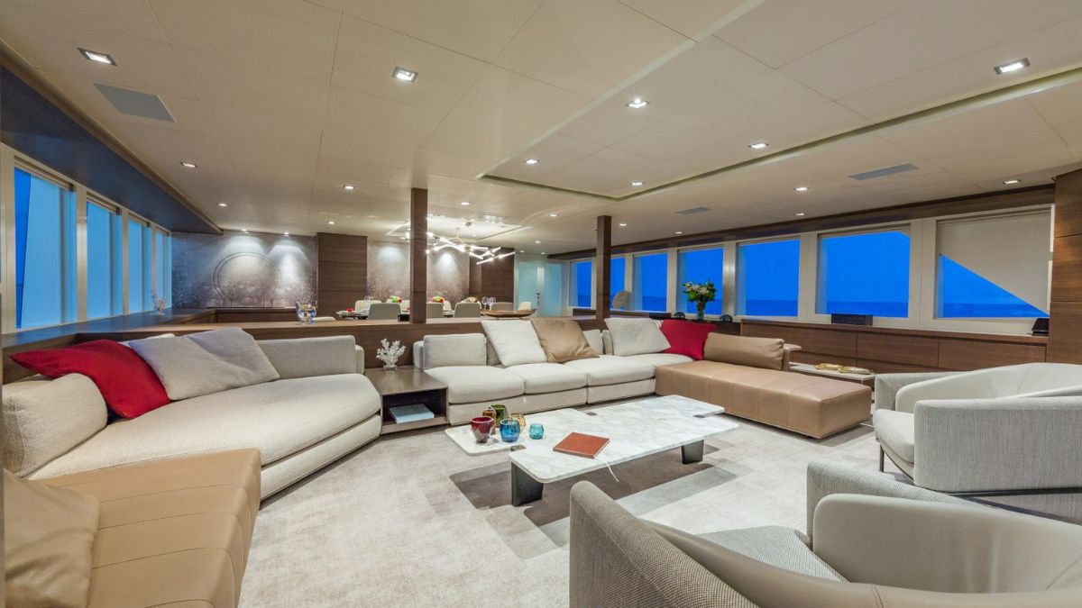 A luxurious yacht interior featuring a spacious lounge area with an L-shaped sectional sofa adorned with red and beige cushions. A glass coffee table sits on a plush carpet, surrounded by two white armchairs. Large windows provide a panoramic view of the ocean and a serene atmosphere.