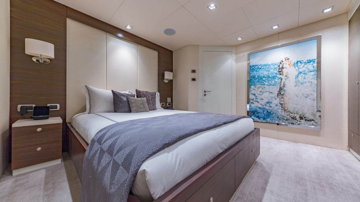 A modern bedroom featuring a large bed with a grey patterned throw, two bedside lamps, and a light wooden decor. A vibrant artwork of a person splashing in water is displayed on the wall. The room has a carpeted floor and subtle lighting accents.