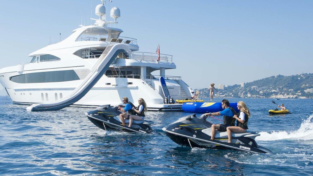 Two jet skis speed across the water, with riders enjoying the sunny day. In the background, a large luxury yacht features a water slide, and several people are engaged in water activities, including lounging and tubing. The scene captures a vibrant day on the water.